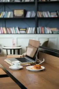 A laptop and croissant on a wooden table in a cozy cafe library setting.